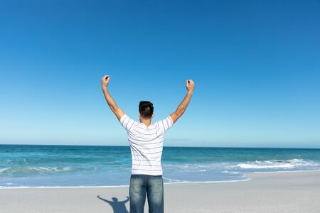 Side view of a Caucasian man standing on the beach with blue sky and sea in the background, raising...