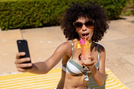 Front view close up of a mixed race woman sitting in the garden by a swimming pool wearing a bikini...