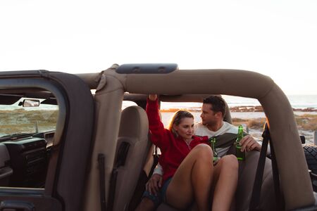 Front view of a Caucasian couple inside an open top car, with sunset on the beach in the background,...