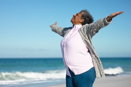 Side view of a senior African American woman standing on the beach with blue sky and sea in the...