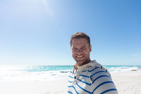 Front view of a Caucasian man standing on the beach with blue sky and sea in the background, smiling to cameraの写真素材
