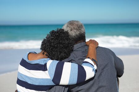 Rear view close up of a senior African American couple sitting on the beach with blue sky and sea in the background, embracingの写真素材