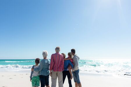 Rear view of a multi-generation Caucasian family standing on the beach with blue sky and sea in the...