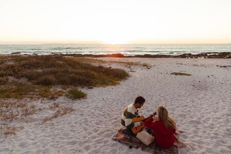 Rear high angle view of a Caucasian couple reclining on the beach, with sunset in the background,...