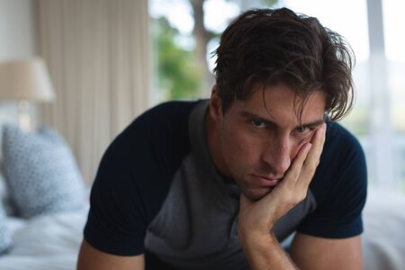 Front view close up of a distressed Caucasian man at home, sitting on his bed, leaning his head on his hand and looking to cameraの写真素材