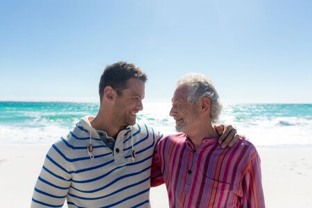 Portrait of a Caucasian man and his father standing on the beach with blue sky and sea in the...