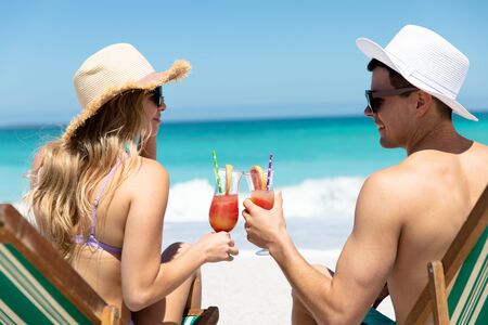Rear view of a Caucasian couple sitting on deckchairs on the beach with blue sky and sea in the background, smiling, looking at each other and making a toastの写真素材