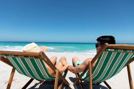 Rear view of a Caucasian couple sitting on deckchairs on the beach with blue sky and sea in the...