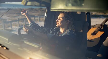 Front view of a Caucasian woman inside an open top car, holding a steering wheel, raising her hands and relaxing in the sun with eyes closedの写真素材