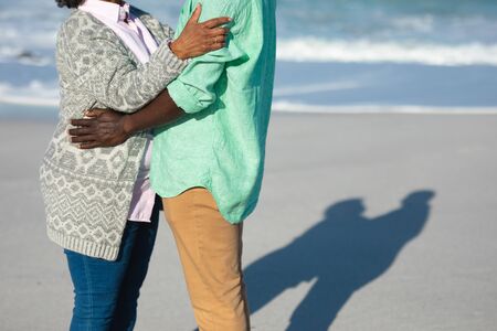 Side view mid section of a senior African American couple standing on the beach with blue sky and sea in the background, embracing の写真素材