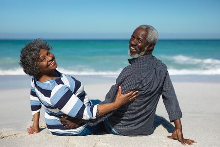 Rear view of a senior African American couple sitting on the beach with blue sky and sea in the background, embracing and turning around, smiling to cameraの写真素材