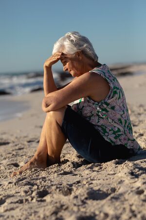 Side view of a senior Caucasian woman at the beach in the sun, sitting on the sand, shielding her eyes, with blue sky in the backgroundの写真素材