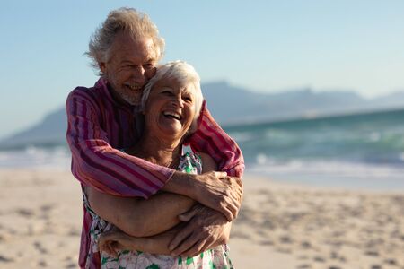 Front view of a senior Caucasian couple at the beach in the sun, embracing and laughing, with blue...