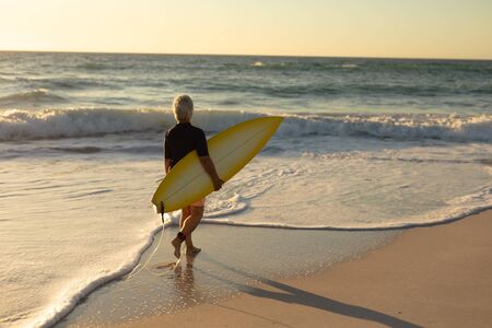 Side view of a senior Caucasian woman at the beach at sunset, walking on the sand carrying a...