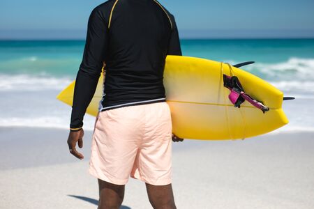 Rear view mid section of a senior African American man on a beach in the sun, carrying a surfboard...