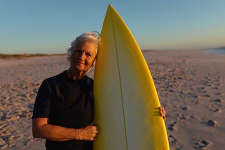 Front view of a senior Caucasian woman at the beach at sunset, standing on the sand holding a surfboard, looking to camera and smilingの写真素材
