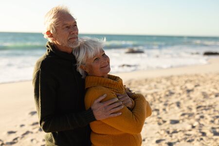 Side view of a senior Caucasian couple at the beach wearing sweaters, embracing and smiling, with...