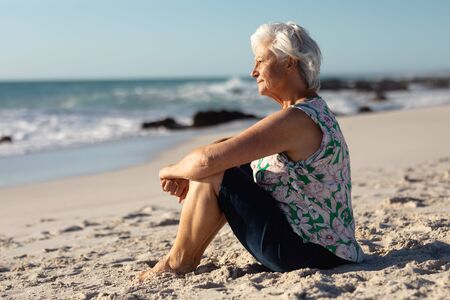 Side view of a senior Caucasian woman at the beach in the sun, sitting on the sand and looking out to sea, with blue sky in the backgroundの写真素材