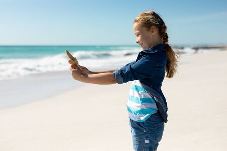 Side view of a Caucasian girl on a sunny beach, holding a starfish in her hand, looking to camera...