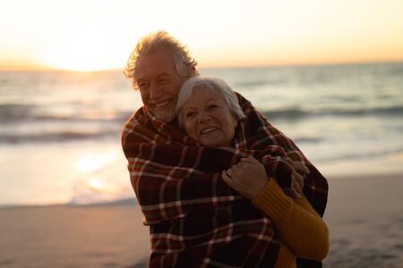 Front view close up of a senior Cacuasian couple standing on the beach at sunset with a blanket over their shoulders, smiling to camera and embracing, with the sea in the backgroundの写真素材
