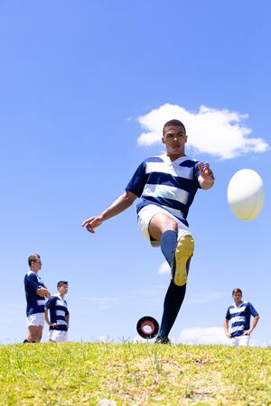 Low angle front view of a mixed race male rugby player kicking a rugby ball, with a group of Caucasian male teammates standing behind him, all wearing a team strip, on a rugby pitch during a match, with blue sky in the backgroundの写真素材
