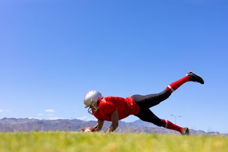 Low angle side view of a mixed race male American football player wearing a team uniform, training at a sports field, diving to the ground holding a football, with blue sky in the backgroundの写真素材