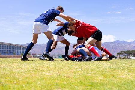 Low angle side view of a mixed race group of male rugby players from two opposing teams wearing team uniforms, in a scrum on a rugby pitch during a match, with blue sky in the backgroundの写真素材