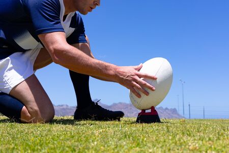 Low angle side view close up of a Caucasian male rugby player wearing a team uniform, training at a sports field, kneeling and placing a rugby ball for a kick, with blue sky in the backgroundの写真素材