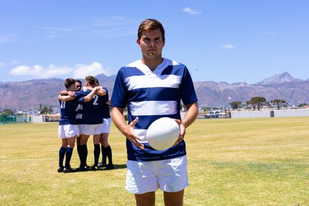 Front view of a Caucasian male rugby player holding the ball, with a mixed race group of male...