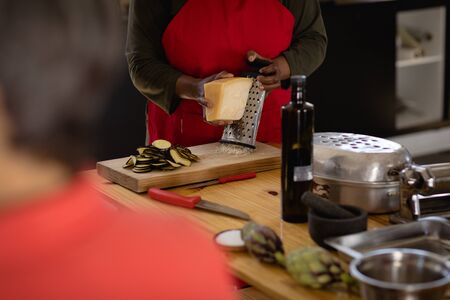 Front view mid section of an Senior African American woman wearing a red apron preparing food at a cookery class, grating cheese with a cheese grater on a wooden board, another cook in the foreground, focus on the backgroundの写真素材