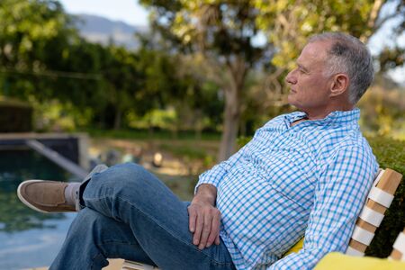 Weekend at home. Side view of a senior Caucasian man in the garden, sitting on a chair relaxing and thinkingの写真素材