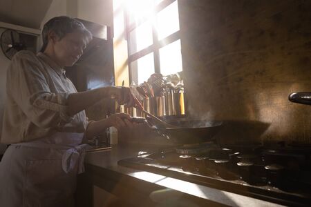 Side view of a Senior Caucasian woman at a cookery class, frying vegetables on a frying pan. Active...