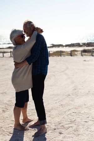 Side view of a senior Caucasian couple sitting at the beach in the sun, embracing and smiling. Weekend beach vacation, lifestyle and leisure.の写真素材