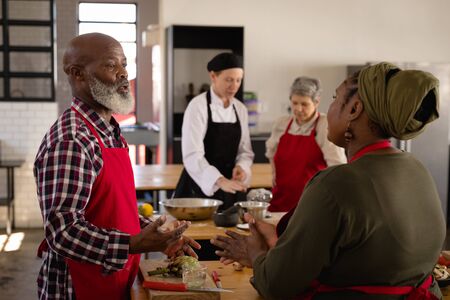 Side view of a multi-ethnic group of Senior adults at a cookery class, an African American couple standing at a wooden table of ingredients talking in the foreground, and a Caucasian woman listening to instructions from a Caucasian female chef wearing chefs whites and a black hat and apron in the backgroundの写真素材