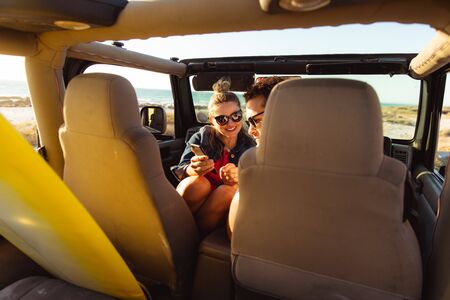 Front view of a Caucasian couple inside an open top car, smiling and using their mobile phone....