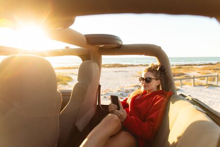 Front view of a Caucasian woman sitting inside an open top car, using her mobile phone and smiling....