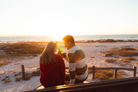 Rear view of a Caucasian couple with sunset on the beach in the background, making a toast. Weekend...