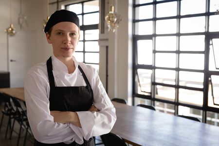 Portrait of a confident Caucasian woman wearing chefs whites and a black apron and hat at a cookery class in a restaurant, standing with arms crossed looking to cameraの写真素材