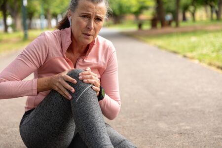 Front view of a mature middle aged Caucasian woman working out in a park, holding her injured knee,...