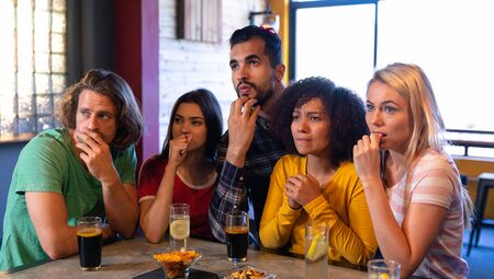 Front view of a group of five multi-ethnic male and female friends at the bar in a pub in suspense during a tense moment while watching a sports game on TV, concentrating and focused in anticipationの写真素材