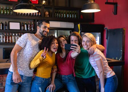 Front view of a group of five multi-ethnic male and female friends at the bar in a pub during the day, one woman holding a smartphone and taking a selfie of them having fun posing for the picture, smiling, laughing and gesturing togetherの写真素材