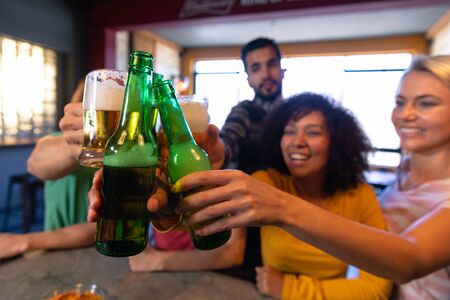 Side view close up of a group of five multi-ethnic male and female friends at the bar in a pub, holding bottles of beer and raisng them to make a toast, smiling and laughing in celebration togetherの写真素材