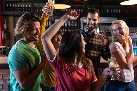 Front view of a group of five multi-ethnic male and female friends at the bar in a pub holding drinks, dancing and having fun and celebrating, two women with arms in the air and all of them smiling and laughing togetherの写真素材