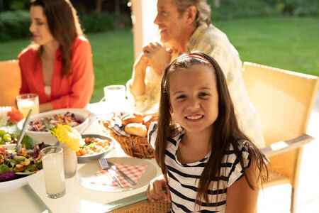 Front view of a young Caucasian girl sitting outside with her family at a dinner table set for a...