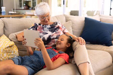 Front view of Caucasian grandmother sitting on a sofa in living room with her granddaughter lying on her lap, using a tablet computer.の写真素材