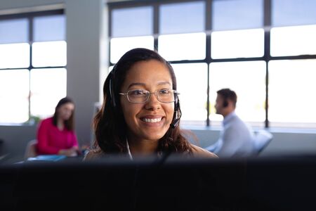 Front view close up of a mixed race woman working as a customer support, wearing smart clothes and headset, using a desktop computer with colleagues working in the background.の写真素材