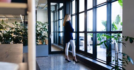 Side view of a Caucasian businesswoman wearing smart clothes, working in a modern office, standing by the window and looking outside.の写真素材