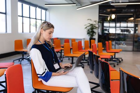 Side view of a Caucasian woman with long blond hair, wearing smart clothes, sitting on a chair in an empty modern meeting room, using a laptop.の写真素材