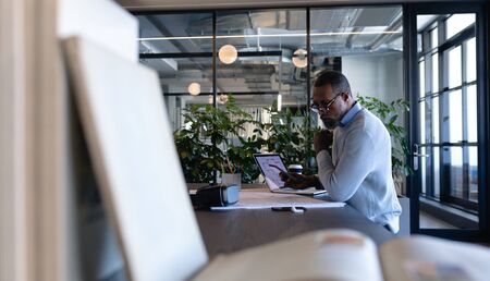 Side view of an African American businessman working in the modern office, sitting by a desk and using his smartphone. Social distancing and self isolation in quarantine lockdownの写真素材