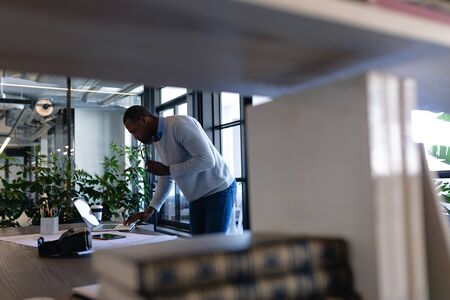 Side view of an African American businessman working in the modern office, standing by a desk, using his laptop and using his smartphone. Social distancing and self isolation in quarantine lockdownの写真素材